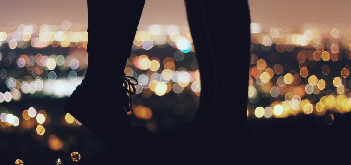 Picture of two silhouette peoples legs / feet kissing in front of a blurred city full of lights.